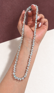 Eveline mixed-shape moissanite necklace in sterling silver with thick rhodium plating, held with very pale milky pink nails against plum and warm paper.